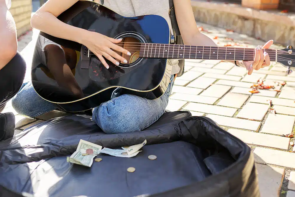 Junge Straßenmusikanten, Nahaufnahme der Hand eines Mädchens, das Akustikgitarre spielt, und Koffer mit Bargeld auf dem Bürgersteig Junge Straßenmusikanten, Nahaufnahme der Hand eines Mädchens, das Akustikgitarre spielt, und Koffer mit Bargeld auf dem Bürgersteig
