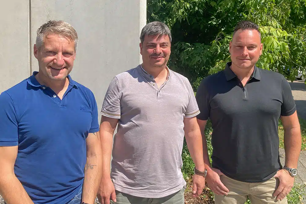 Auf dem Bild sind drei Personen zu sehen. Von links nach rechts: Christian Huth von Apex International, Markus Musoke von DEBATIN und Meikel Maier von Doneck Euroflex S.A.. Sie stehen nebeneinander draußen und lächeln in die Kamera.