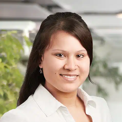 Nahaufnahme von Alexandra Böhmer, sie ist Teil des Einkauf / Purchasing Department bei Debatin. Man sieht eine lächelnde Frau mit langen, dunklen Haaren und einem Nasenpiercing. Sie trägt eine weiße Bluse, und im unscharfen Hintergrund sind grüne Pflanzen und helle Strukturen zu erkennen.