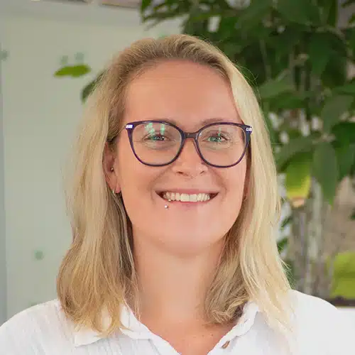 Porträt von Denise Raupp, Mitarbeiterin im Einkauf / Purchasing Department bei Debatin. Sie lächelt freundlich und trägt eine Brille sowie ein Lippenpiercing. Ihr blondes Haar fällt schulterlang, und sie trägt ein weißes Oberteil vor einem unscharfen grünen Hintergrund.