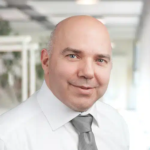 Kopf- und Schulteraufnahme von Helmut Roth, der im Einkauf / Purchasing Department bei Debatin tätig ist. Er blickt direkt in die Kamera mit einem leichten Lächeln. Der Mann mit Glatze trägt ein weißes Hemd und eine graue Krawatte. Der Hintergrund ist unscharf und deutet auf eine Büroumgebung hin.