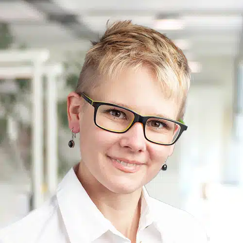 Melanie Kost, eine Mitarbeiterin des Versand / Shipping Department bei Debatin, blickt mit einem zugewandten Lächeln in die Kamera. Ihre kurzen, platinblonden Haare und die stylische Brille mit neon-grünen Akzenten verleihen ihr eine individuelle Note.