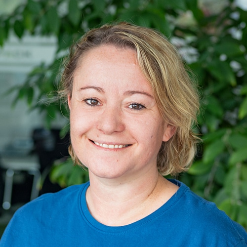 Nahaufnahme von Michaela Breßer, sie ist als Vertriebsberaterin / Customer Service bei Debatin tätig. Man sieht eine lächelnde Frau mit kurzen, blonden Haaren, die ein blaues T-Shirt trägt. Im Hintergrund sind grüne Pflanzen verschwommen zu erkennen.