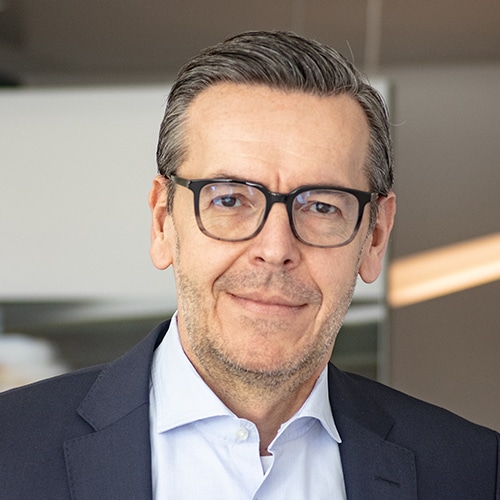 Porträtaufnahme von Oliver Kosalla, dem Leiter Einkauf / Head of Purchasing bei Debatin. Er trägt eine moderne Brille, ein dunkelblaues Sakko über einem hellblauen Hemd und blickt direkt in die Kamera. Sein Gesicht zeigt einen gepflegten Bartansatz und grau meliertes Haar.