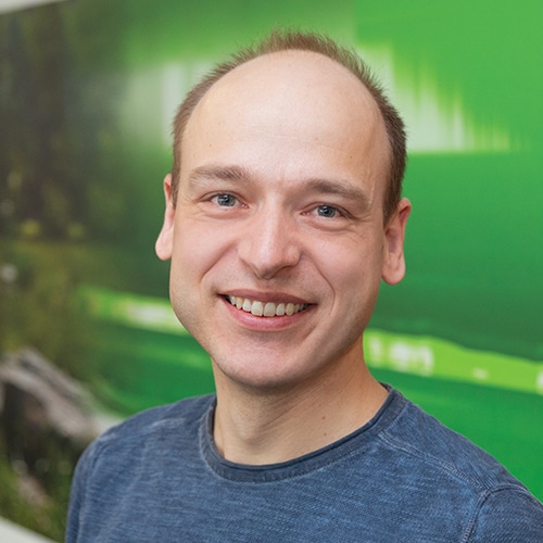 Porträt von Oliver Tröber, Teil des Entwicklung / Development Teams bei Debatin. Er lächelt freundlich in die Kamera, trägt ein schlichtes blaues Oberteil und hat kurze, helle Haare. Der Hintergrund zeigt eine unscharfe grüne Fläche.