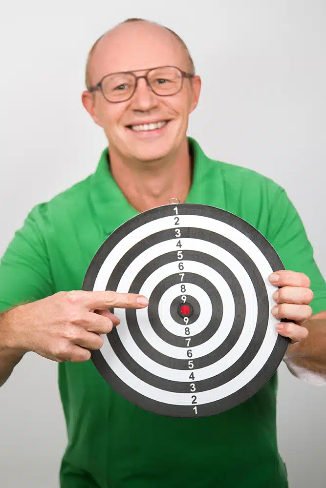 DEBATIN_Produktsparten_Headerbild_DEBADEINS_Gerald Frank_Zielscheibe Gerald Frank trägt ein grünes Shirt und hält eine bunte Dartscheibe hoch.