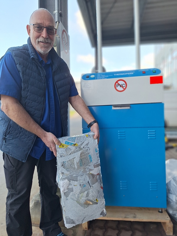 Jürgen Heß, Leitung Materialwirtschaft bei der Bioscientia Healthcare GmbH, steht neben der blauen Kunststoff-Recyclingpresse von DEBATIN. Er hält einen gepressten Block aus recyceltem Kunststoffmaterial in den Händen. Im Hintergrund ist ein überdachter Außenbereich eines Industriegebäudes zu sehen.