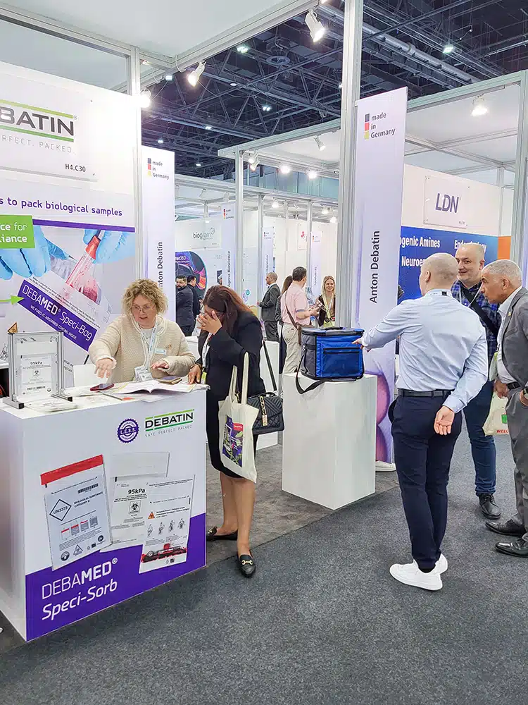 Mehrere Personen stehen an einem Messestand mit der Beschriftung „Anton Debatin“. Eine Person zeigt auf Unterlagen und Produktbeispiele für „DEBAMED® Speci-Sorb“, während andere Personen zuhören oder sich umsehen.