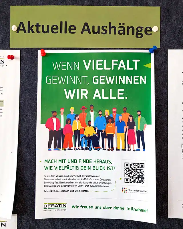 Ein Aushang hängt mit farbigen Pinnnadeln an einer Filzwand unter der Überschrift „Aktuelle Aushänge“. Das Poster bewirbt den Deutschen Diversity-Tag mit einer Illustration verschiedener Personen, einem QR-Code und dem Slogan „Wenn Vielfalt gewinnt, gewinnen wir alle.“